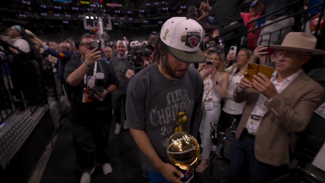 Jalen Brunson is slowly hobbling to the locker room after the 2026 NBA Finals championship ceremony holding the Finals MVP TrophyHis wife orange stripe can be seen alongside him